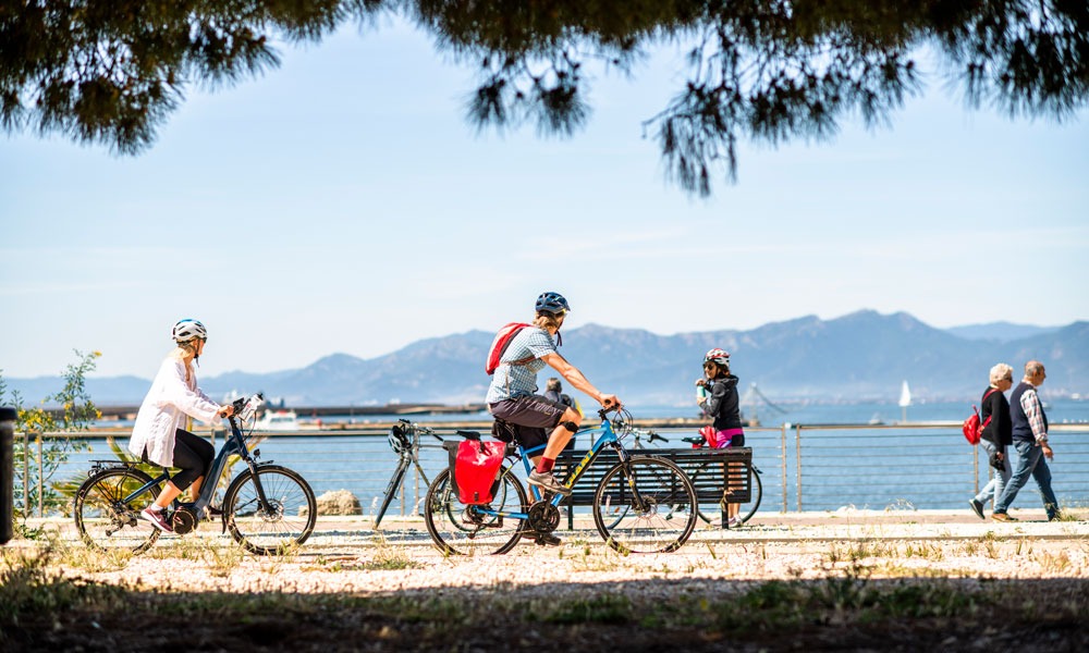 Two cyclists cruise along a promenade with sea and mountains behind