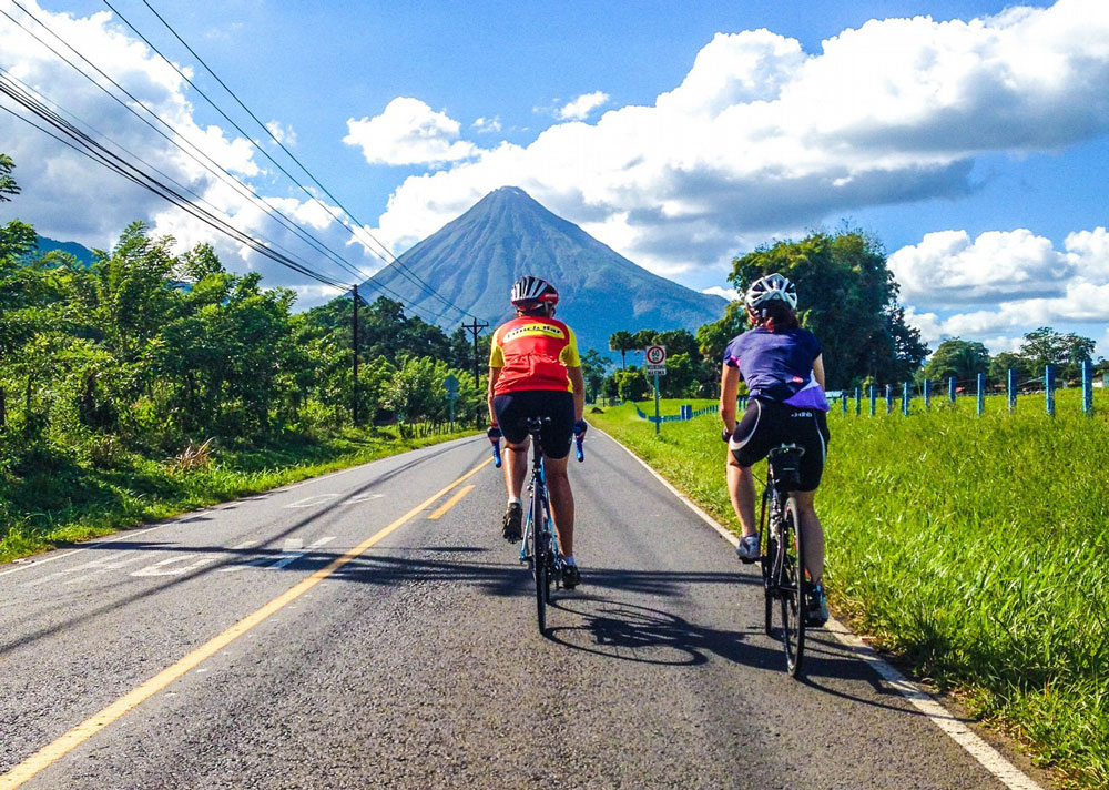 Two cyclists ride on an empty road with a mountain in the distance