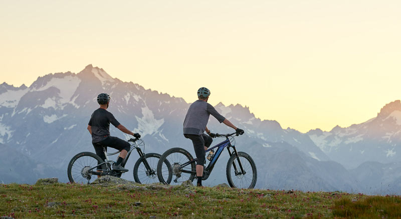 Two people sit on electric mountain bikes at the top of a mountain looking across at a sunset mountain view