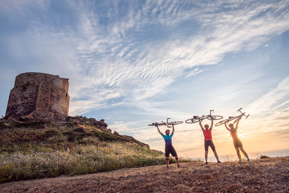 Three cyclists smile holding their bikes above their head. with the sunset and coast behind