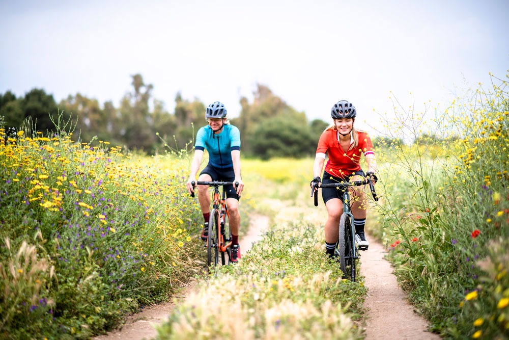 Two people smile as they ride side by side along a double track through a field of flowers