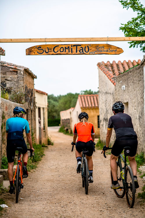 Three cyclists ride into an old village. The sign reads Su Comitau San Salvatore