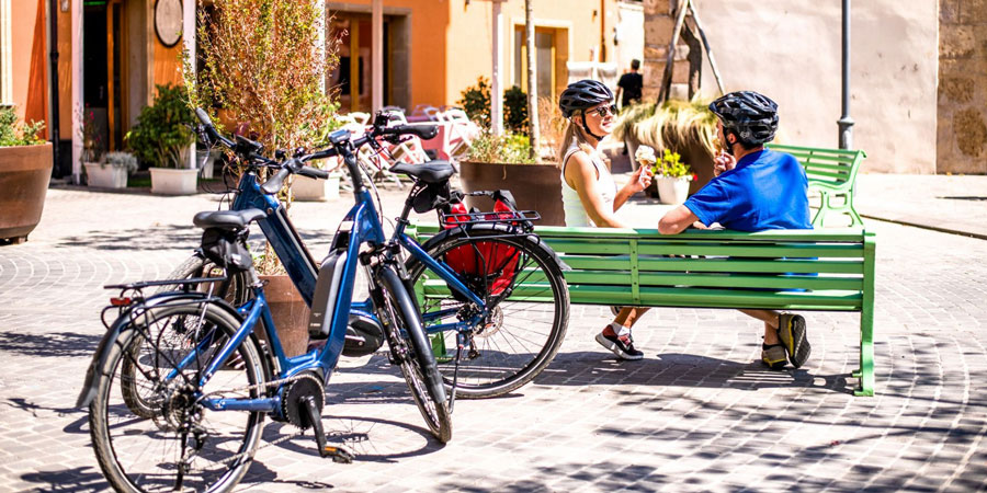 Two people sit on a sunny bench with e bikes behind them