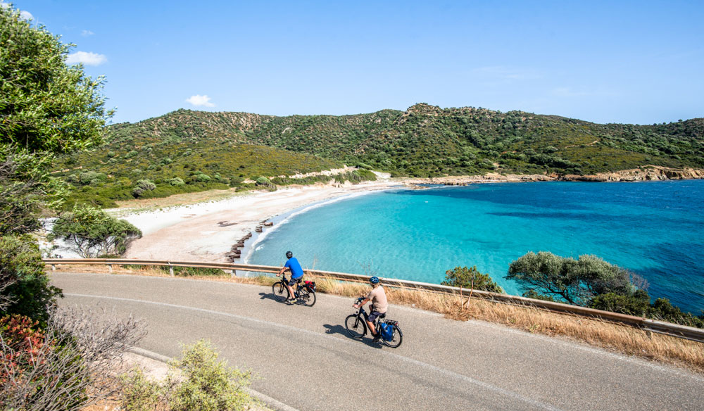 Two people ride ebikes alongside a turquoise ocean in Sardinia