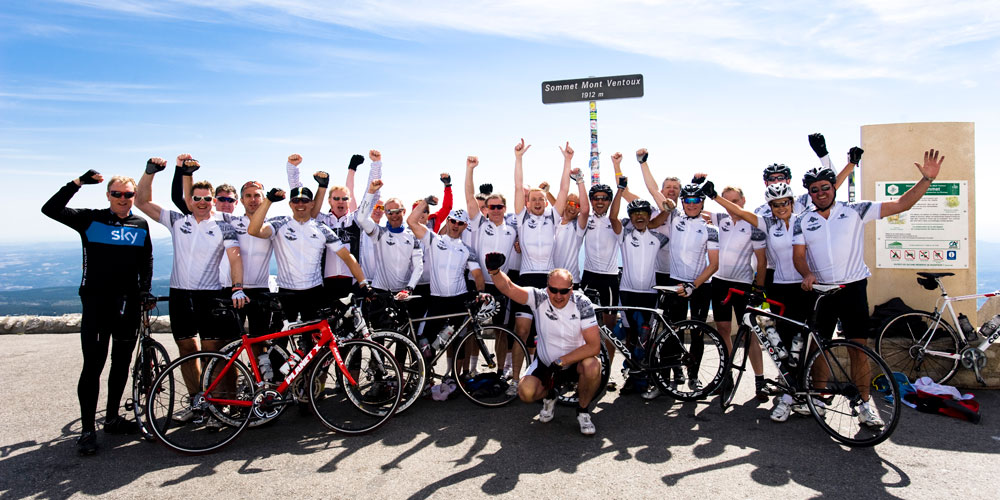 A large group of cyclists throw hands in the air at the summit of Mont Ventoux