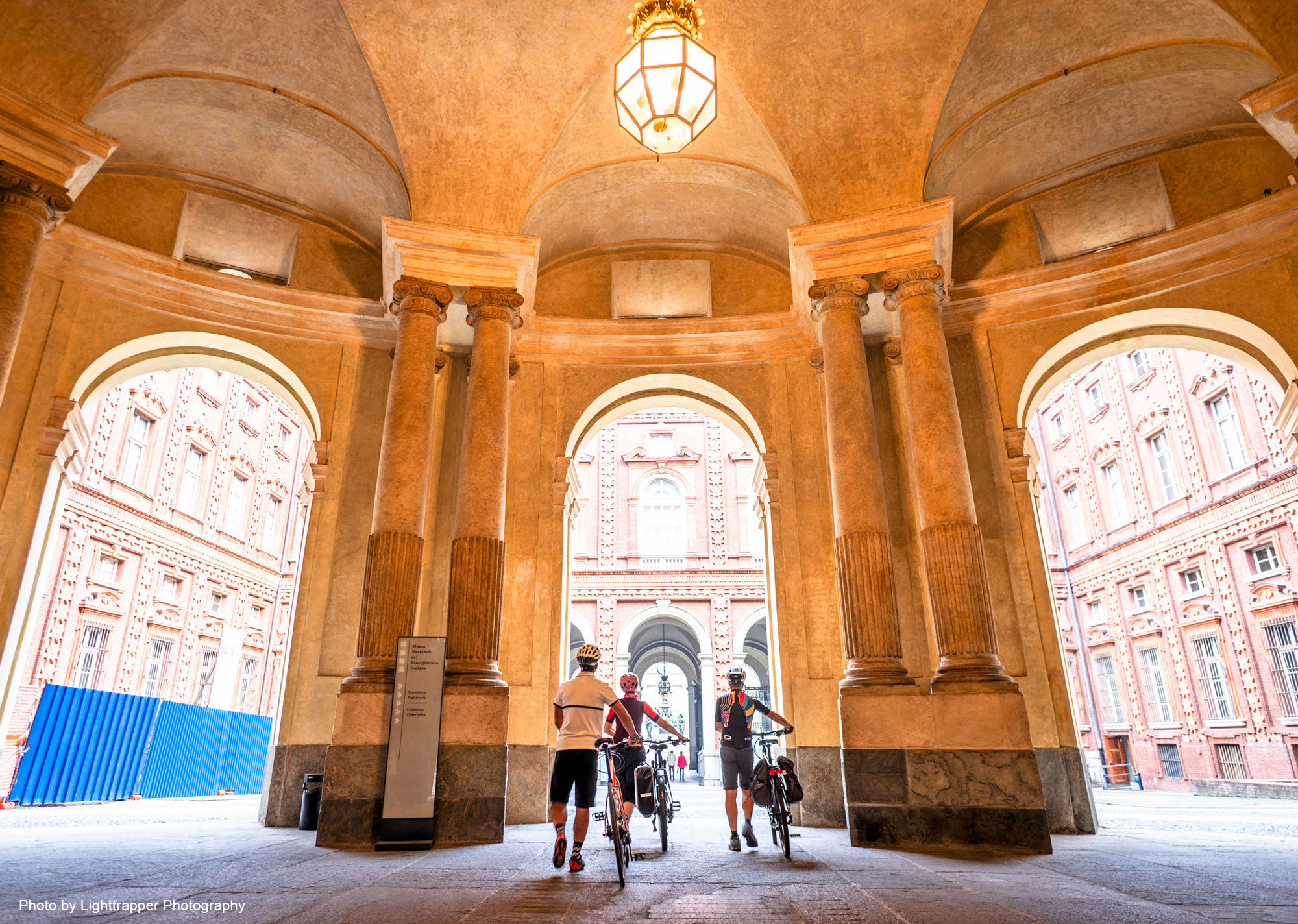 Cyclists wheel their bikes through big arches in a grand building
