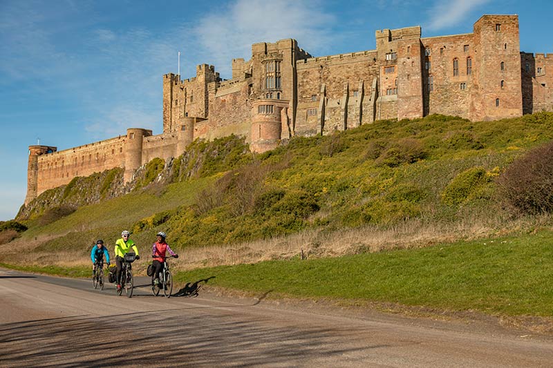 Three cyclists riding past Bamburgh Castle in Northumberland